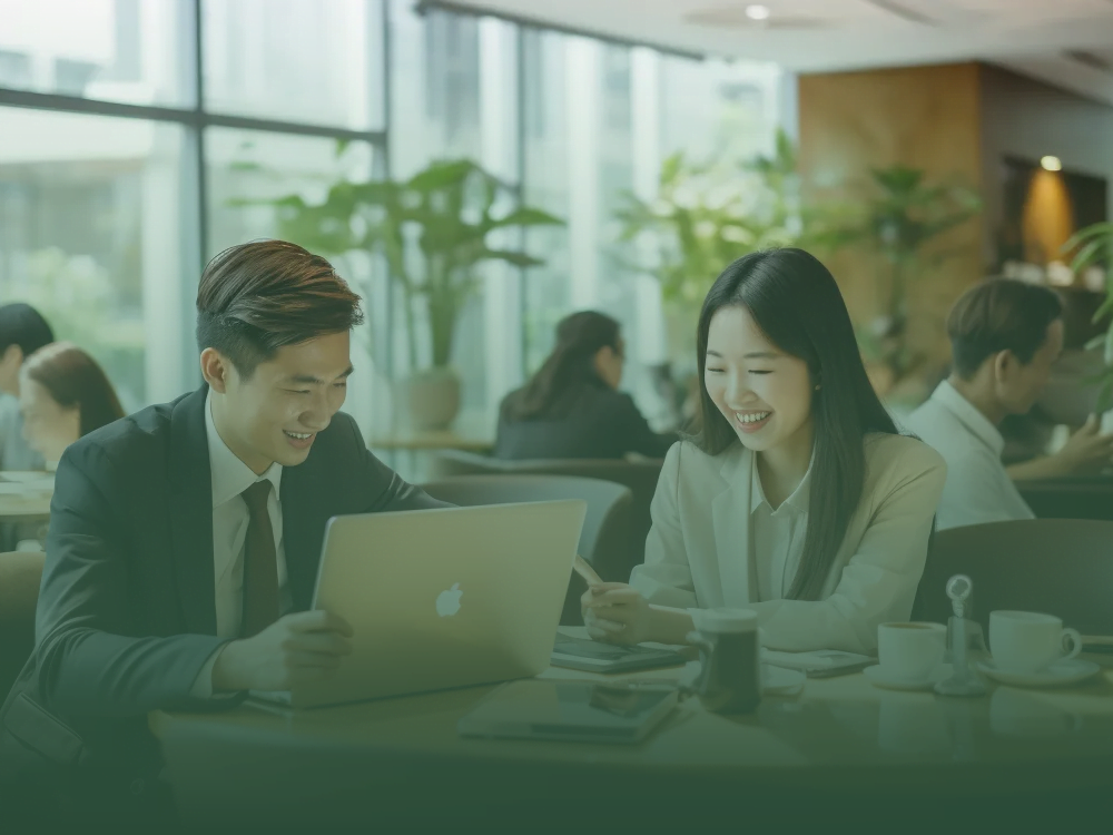 Two professionals smiling while discussing marketing strategies on a laptop in a lively office. Business marketing fundamentals
